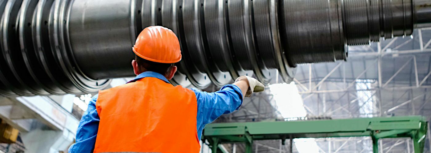 Ein Mann in einer Industriehalle, er trägt Sicherheitskleidung und dirigiert ein großes Maschinenteil an einem Kran.