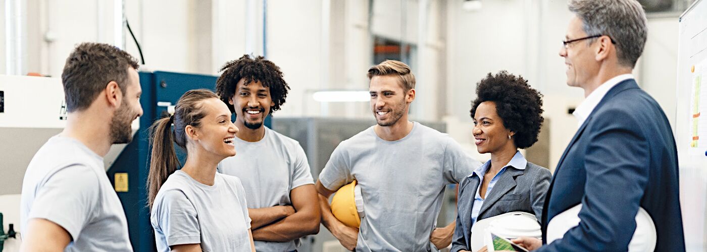 Eine Gruppe von vier Kolleginnen unterhalten sich in einer Fabrikhalle mit der Betriebsleitung