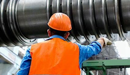 Ein Mann in einer Industriehalle, er trägt Sicherheitskleidung und dirigiert ein großes Maschinenteil an einem Kran.